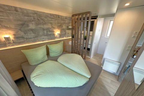 Modern bedroom in a tiny house with green bedding, wooden divider, soft lighting, and bathroom view.