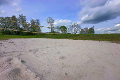 Outdoor sand volleyball court surrounded by grass and trees near a tiny house under a blue sky.