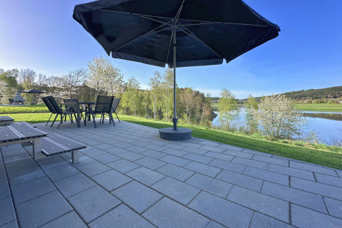 Patio with umbrella and garden furniture overlooking a lake at SEEBLICK Tiny Chalet Holiday Village in Germany.