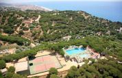 Vue aérienne d’un site de glamping avec piscines, courts de tennis, arbres et vue sur mer près d’une ville côtière.