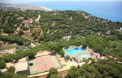 Aerial view of a glamping site with swimming pools, tennis courts, lush trees, and scenic sea views nearby.