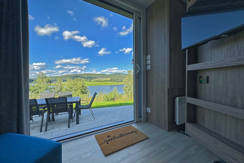 View from a tiny house through a glass door to patio with table, chairs, lake, and hills in the background.