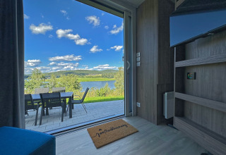 Vue d'une tiny house à travers une porte vitrée sur une terrasse avec table, chaises, lac et collines.