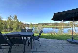Patio with dining table, chairs, and sun umbrella overlooking a lake and hills at a tiny house in Germany.