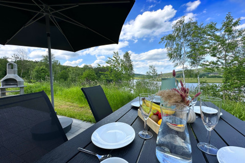 Udendørs spiseområde med sort bord, hvidt service, frugt og udsigt over sø og skov ved SEEBLICK Tiny Chalet.