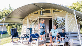 Gezin ontspant buiten voor een safaritent en geniet samen van koffie en snacks op het houten terras.