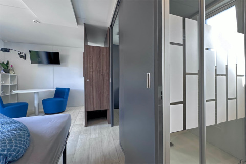 Interior de tiny house moderno con cama, área de estar, mesa, TV y ducha con cristales esmerilados en Alemania.