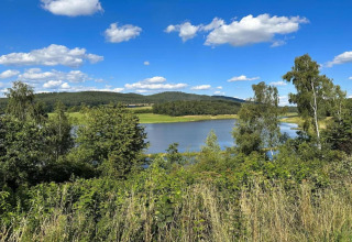 View from a tiny house overlooking a peaceful lake, surrounded by green trees, hills, and blue sky.