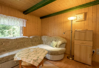 Gemütliches Wohnzimmer in einer Holzhütte mit Ecksofa, Stehlampe, kleinem Tisch und Waldblick.