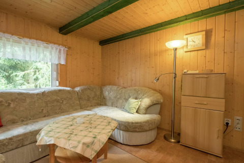 Gezellige woonkamer in blokhut met hoekbank, staande lamp, klein tafeltje en raam met boszicht.
