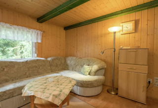 Gezellige woonkamer in blokhut met hoekbank, staande lamp, klein tafeltje en raam met boszicht.