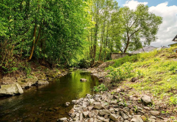 Bach neben einem Ferienhaus mit Parkplatz in Deutschland, umgeben von Bäumen und Natur unter blauem Himmel.