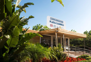 Entrada al Camping & Bungalow Park Platja Cambrils rodeada de plantas tropicales y ambiente soleado.