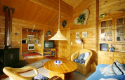 Cozy chalet living room with wooden walls, wicker chairs, wood stove, TV, and an open kitchen view.