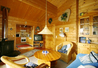 Gezellige chalet woonkamer met houten wanden, rotan stoelen, houtkachel, tv en open keuken erachter.