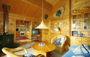 Gezellige chalet woonkamer met houten wanden, rotan stoelen, houtkachel, tv en open keuken erachter.