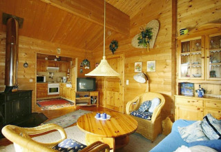 Salon de chalet chaleureux avec murs en bois, fauteuils en osier, poêle, télévision et cuisine ouverte.