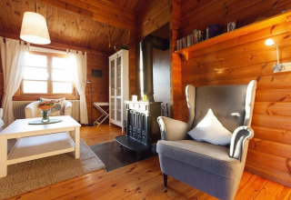 Salon de chalet chaleureux avec murs en bois, poêle, fauteuil gris, table blanche et grande fenêtre.