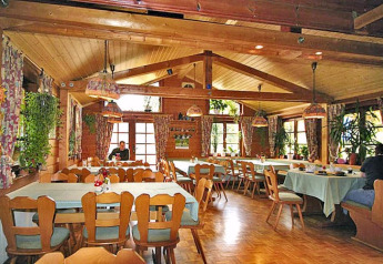 Knus chalet-interieur met houten balken, bloemen gordijnen, veel planten en tafels met tafelkleden.