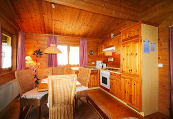 Cozy chalet kitchen and dining area with wooden walls, wicker chairs, and sunlight through purple curtains.