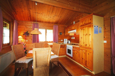Cozy chalet kitchen and dining area with wooden walls, wicker chairs, and sunlight through purple curtains.