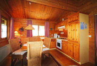 Cocina y comedor de chalet con muebles de mimbre, paredes de madera y cortinas lilas iluminadas por el sol.