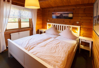 Cozy chalet bedroom with wooden walls, a white double bed, soft lighting, and bright window curtains.