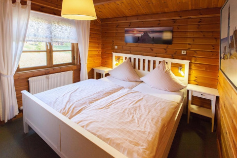 Cozy chalet bedroom with wooden walls, a white double bed, soft lighting, and bright window curtains.