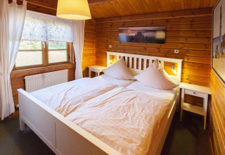 Cozy chalet bedroom with wooden walls, a white double bed, soft lighting, and bright window curtains.
