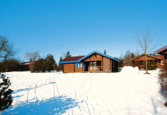 A wooden chalet surrounded by snow and trees under a clear blue sky, with more chalets in the background.