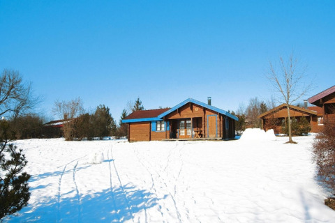 Un chalet de madera rodeado de nieve y árboles bajo un cielo azul claro, con más chalets al fondo.