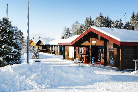 Wooden chalets covered in snow in a peaceful winter setting, surrounded by pine trees and clear blue sky.