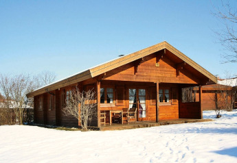 Wooden chalet with covered porch and outdoor furniture, surrounded by snow and leafless trees in winter.