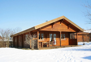 Wooden chalet with covered porch and outdoor furniture, surrounded by snow and leafless trees in winter.