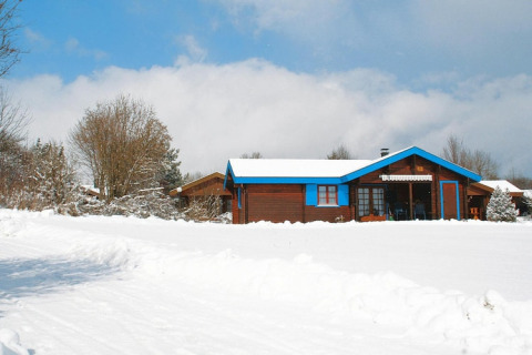 Wooden chalet with blue trim in a snowy landscape, surrounded by snow-covered trees beneath a cloudy sky.