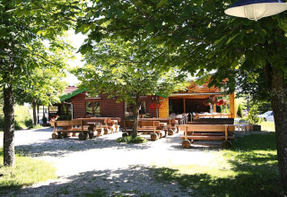 Wooden benches and tables under leafy trees outside a cozy chalet in bright, sunny weather.