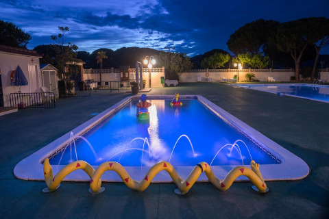Beleuchteter Pool mit Wasserspielen und Spielfiguren auf einem Glampingplatz bei Abenddämmerung.
