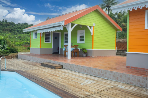 Bungalow colorido con terraza y piscina en medio del campo verde, bajo un cielo azul brillante.
