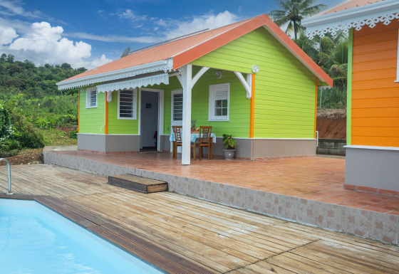 Bungalow colorido con terraza y piscina en medio del campo verde, bajo un cielo azul brillante.
