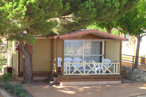 Holzbungalow im Camping Rocagrossa für 5 Personen, Terrasse mit Stühlen und großen schattigen Bäumen.