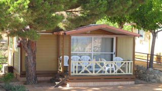 Træbungalow hos Camping Rocagrossa med plads til 5 personer, terrasse med stole under fyrretræer.