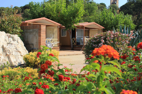 Blomstrende have foran bungalow på Camping Rocagrossa i Spanien, omgivet af træer og farverige blomster.