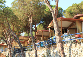 Cabins nestled among pine trees at Camping Rocagrossa Bungalow in Spain, with stone wall and blue railing.