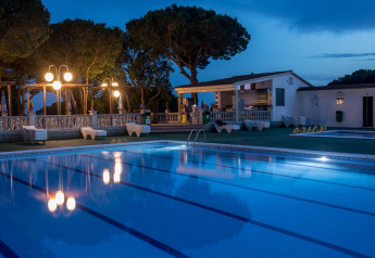 Oplyst swimmingpool ved skumring i et glamping-opholdssted med liggestole, træer og et udendørs opholdsområde.