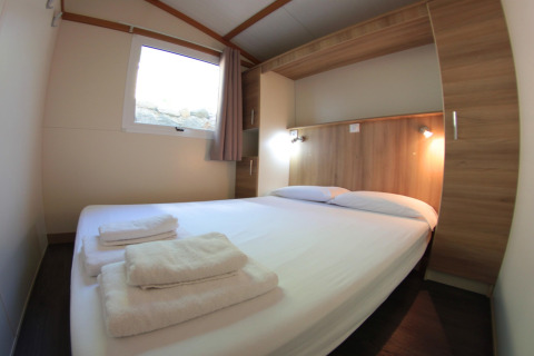 Komfortabelt soveværelse med dobbeltseng, træpaneler og friske håndklæder på Camping Rocagrossa Bungalow i Spanien.