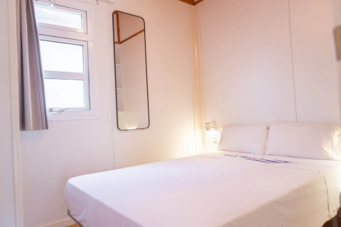 Lichte slaapkamer in Camping del Mar Bungalow Mediterrani, Spanje, met tweepersoonsbed, raam en spiegel.