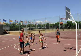 Kinderen en volwassenen spelen basketbal op een buitenveld bij een glampingaccommodatie, met vlaggen ernaast.