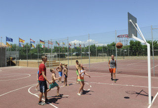 Kinder und Erwachsene spielen Basketball auf einem Outdoorplatz in einer Glamping-Unterkunft mit Flaggen.