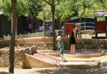 Een kind en een vrouw spelen minigolf onder bomen op een glamping met een speeltuin op de achtergrond.