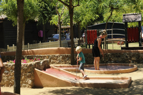 Un enfant et une femme jouent au mini-golf sous les arbres dans un hébergement glamping avec aire de jeux.